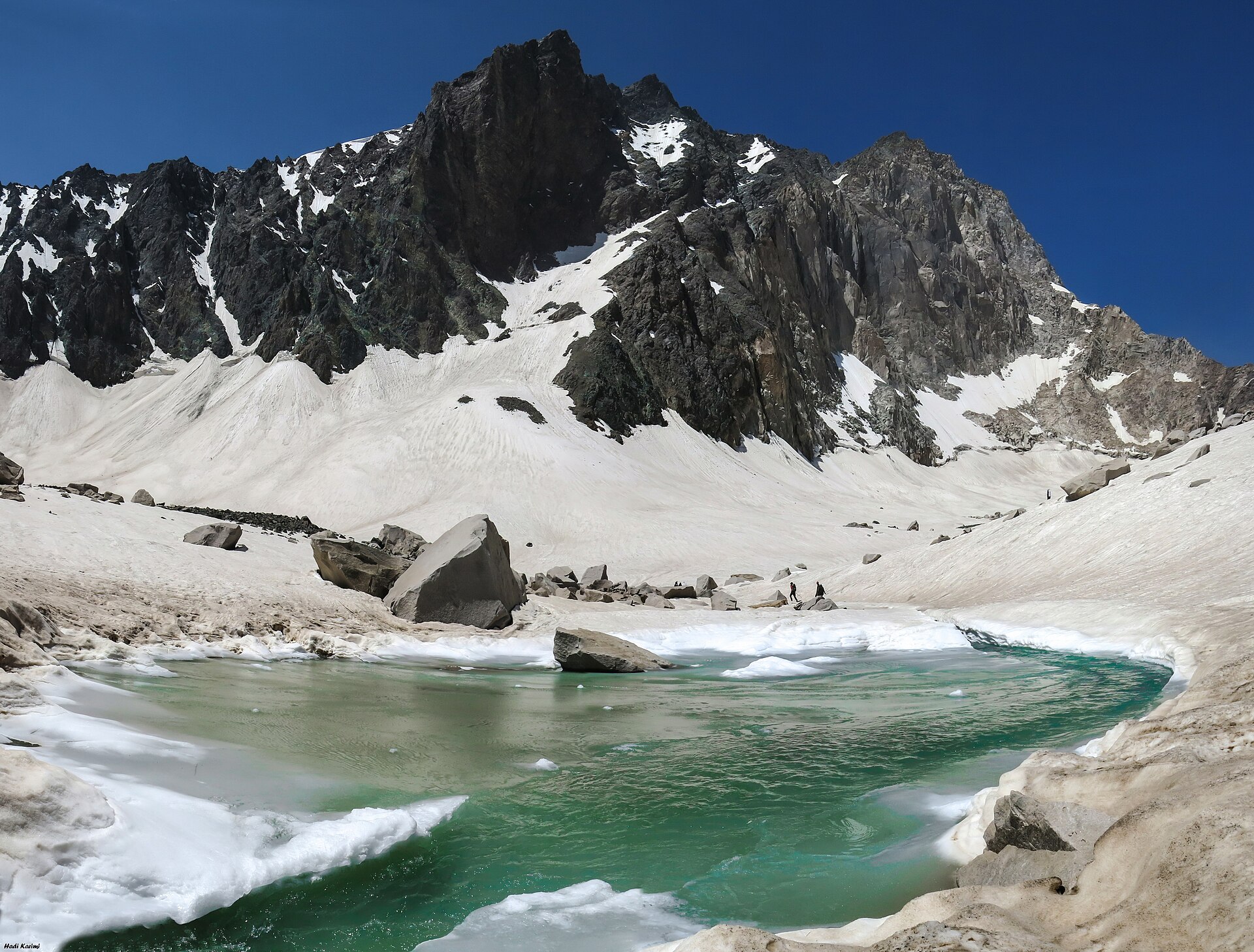 Alam Kuh's rocky north face above snowfields and a turquoise glacial lake